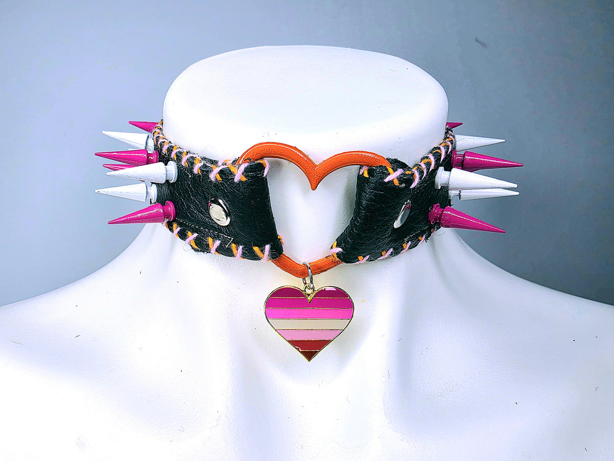 A gothic-style spiked collar necklace displayed on a white mannequin bust. The collar features black faux leather panels, white and pink spikes, a bright orange heart-shaped center, and a pink and orange striped heart pendant.