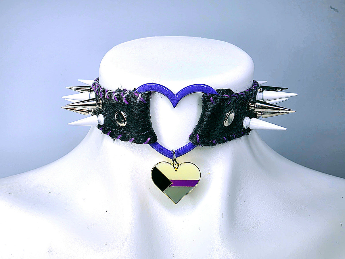 A black leather choker collar with silver metal spikes, purple stitching, and a purple heart-shaped cutout, displayed on a white mannequin bust. A dangling heart charm features the black, purple, and gret colors of the Demisexual flag.