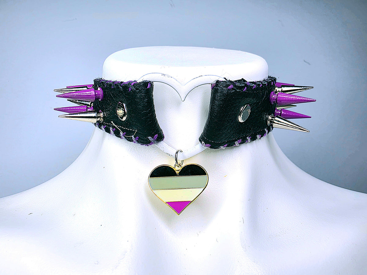 A black leather spiked collar necklace displayed on a white mannequin bust. The collar features multiple sharp silver metal spikes, purple leather trim, and a dangling heart-shaped charm with horizontal stripes in black, olive green, yellow, and purple.