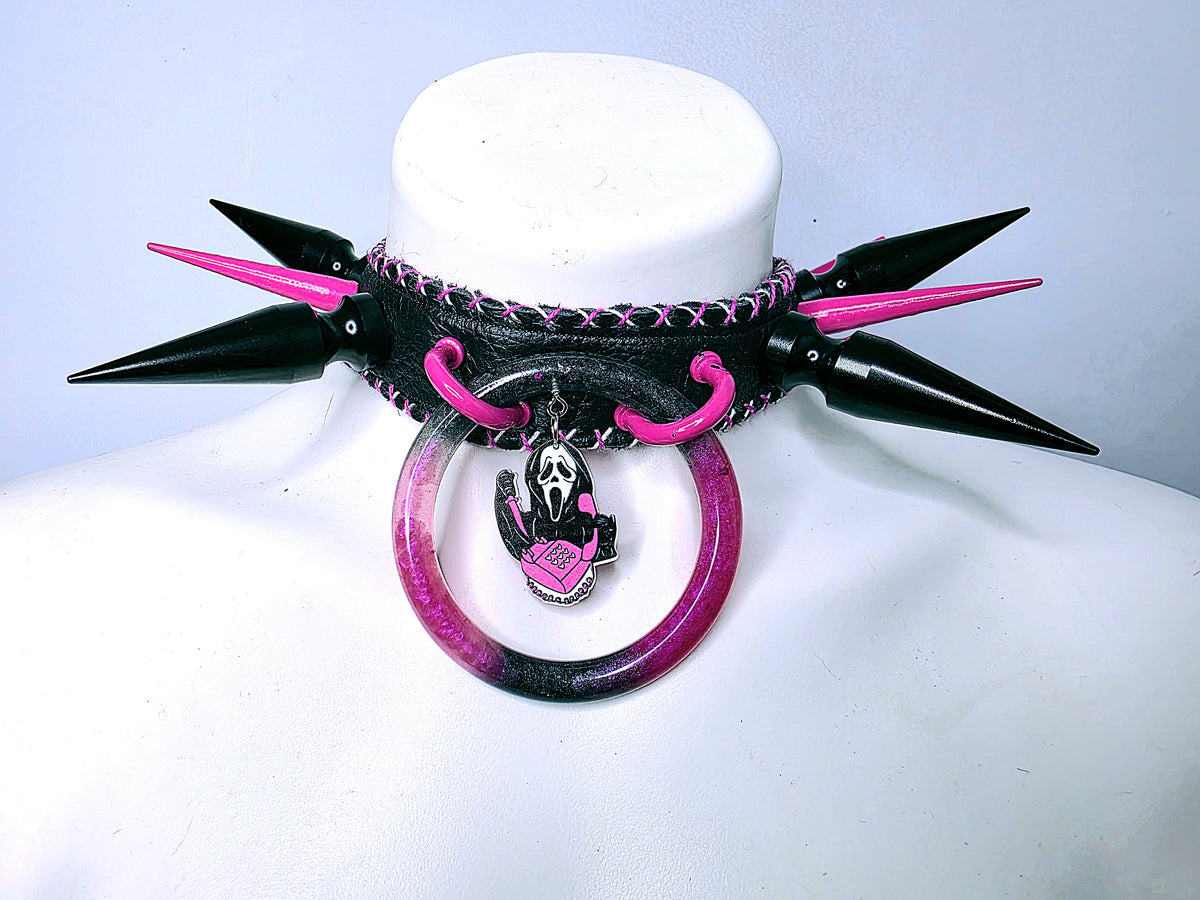A gothic-style spiked collar displayed on a white mannequin bust. The collar features black conical spikes with hot pink tips, a black leather strap with pink stitching, a pink and black circular hoop, and a small metal charm depicting a stylized Ghostface face with a pink heart.