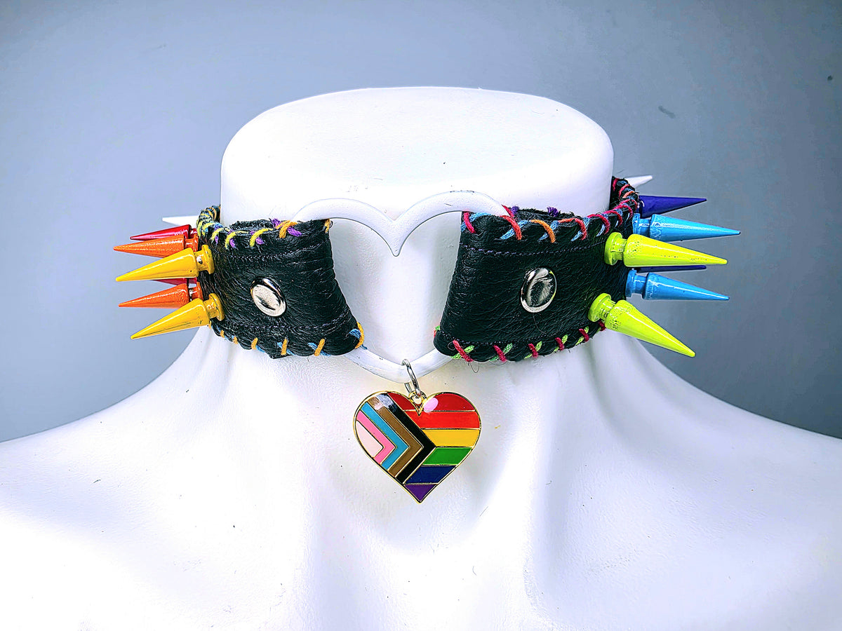 A black leather spiked choker collar displayed on a white mannequin bust. The collar features multiple colorful wooden spikes in yellow, orange, red, blue, green, and purple, along with a rainbow-striped heart-shaped charm.